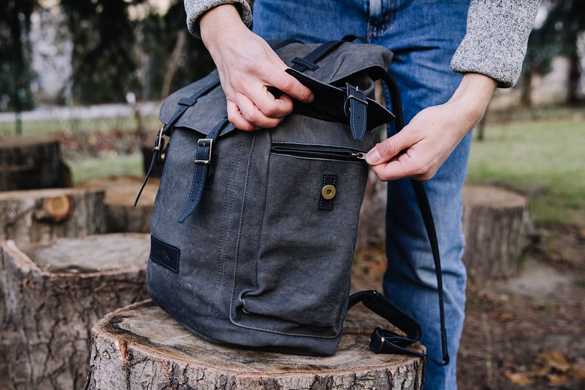Ausgestattet mit Seitentaschen, Rückenpolsterung und viel Stauraum steht der graue Rucksack auf einem Holzstück im Freien.