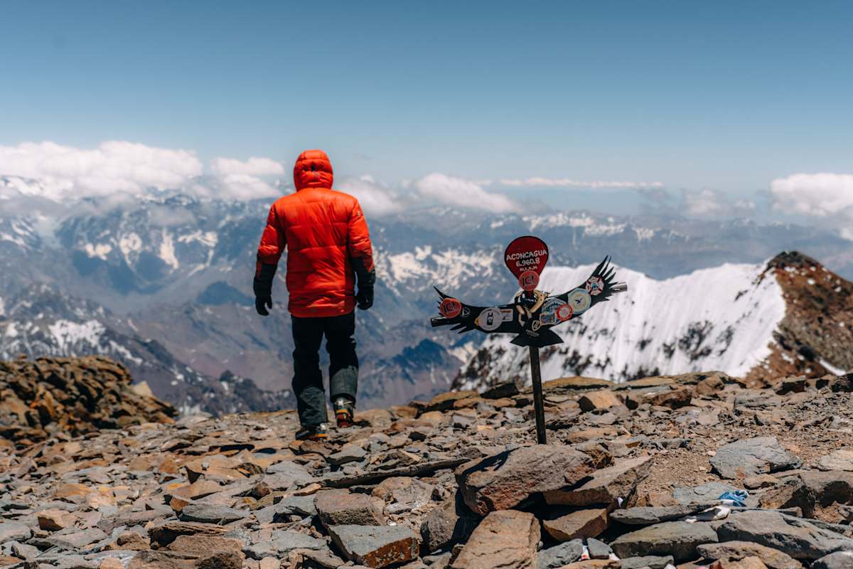 Person in roter Daunenjacke steht am Gipfel des Aconcaguas mit Blick auf die umliegende Bergwelte