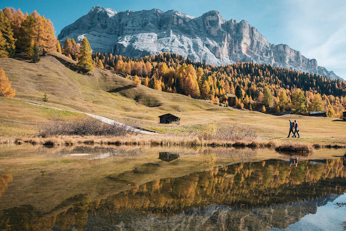 Zwei Wanderer gehen an einem See vorbei, in dem sich die Dolomiten spiegeln.
