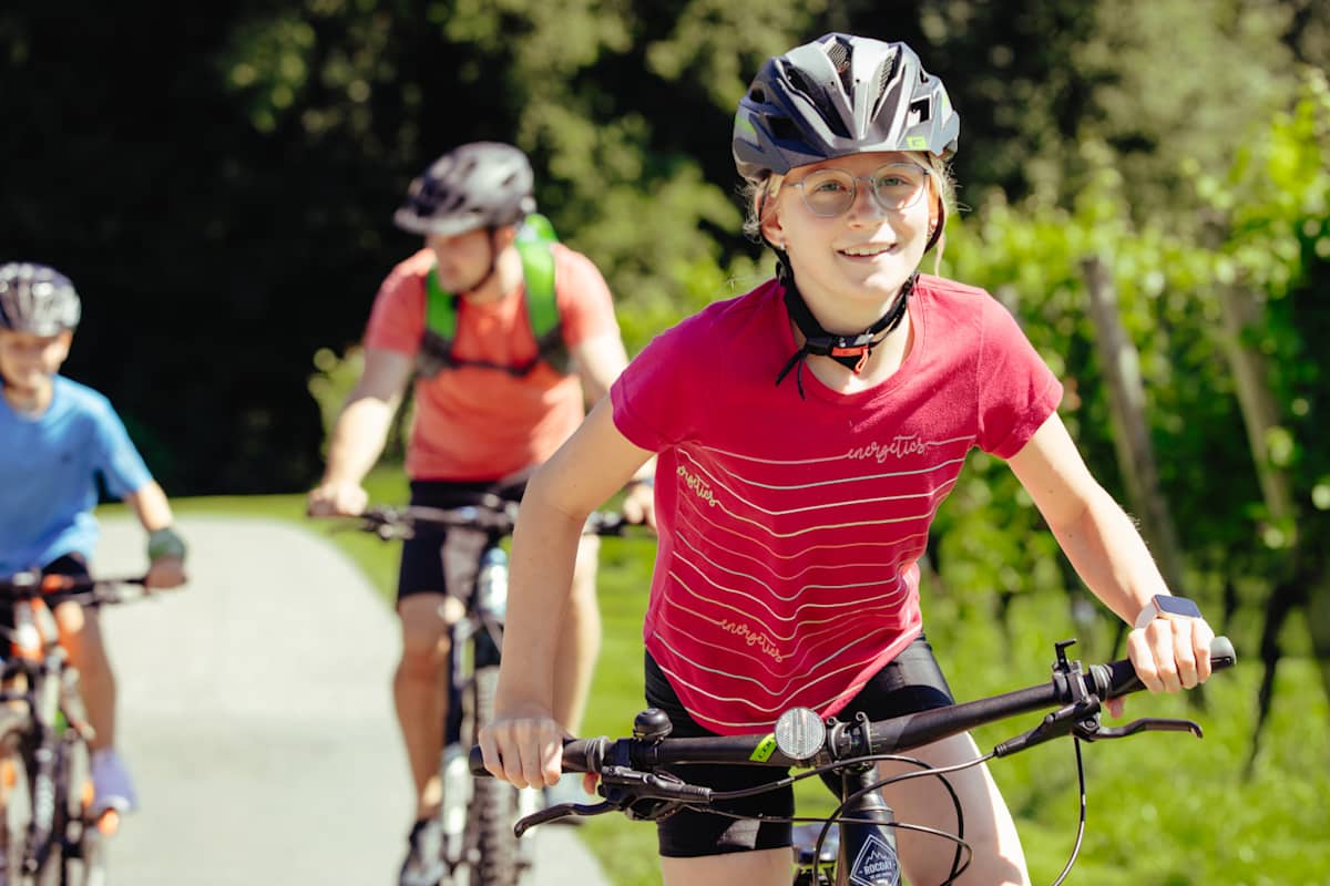 Ein junges Mädchen mit glücklichem Gesicht radelt an Weinbergen vorbei. Im Hintergrund ihr Vater und ihr Bruder.
