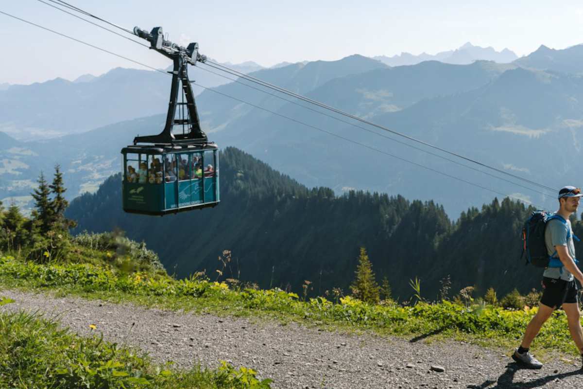 Ein Wanderer geht über eine Forststraße am Walmendinger Horn, im Hintergrund eine Gondel und Berge.
