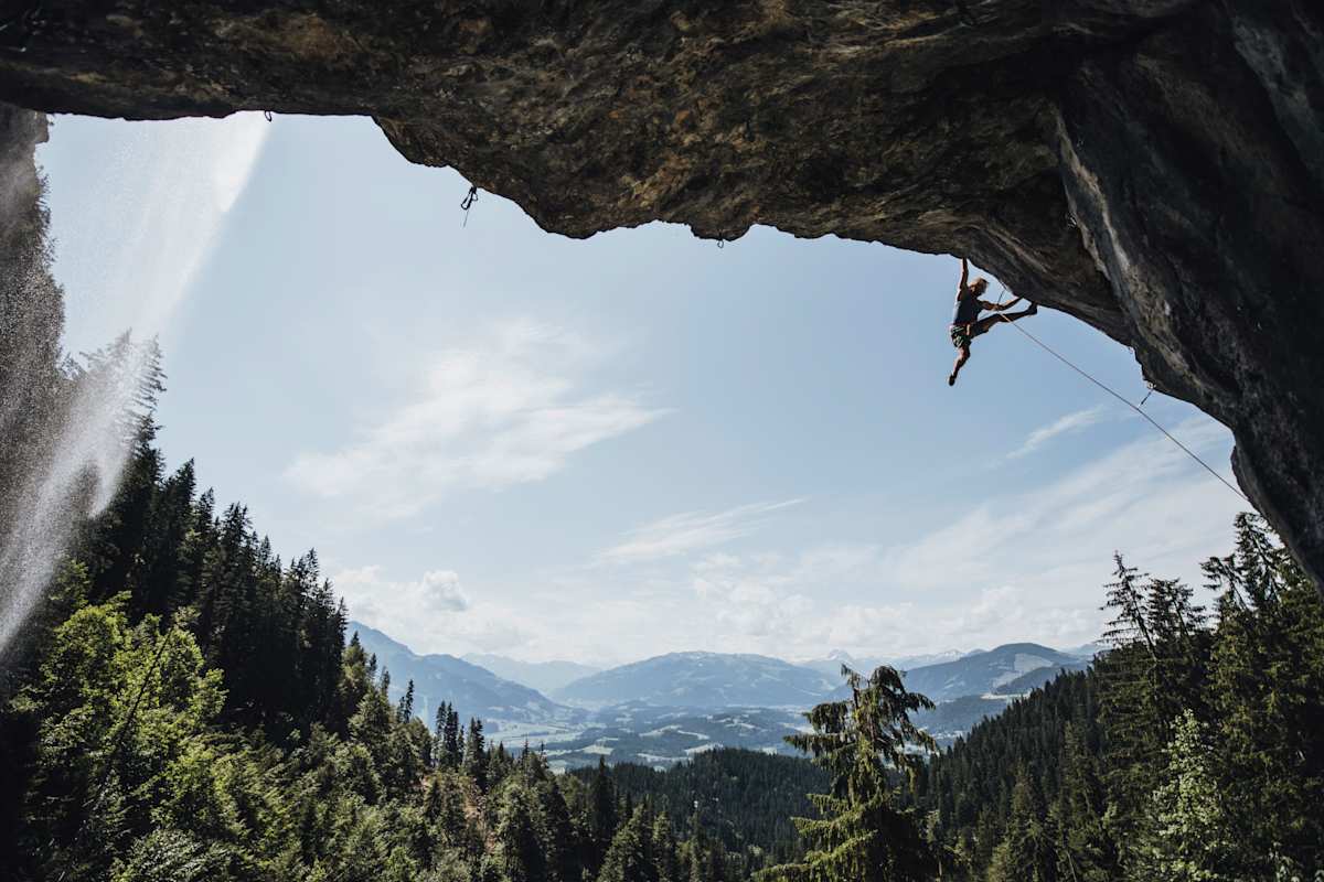 Im Vordergrund: ein Kletterer im Überhang und ein Wasserfall. Im Hintergrund: eine Berglandschaft.