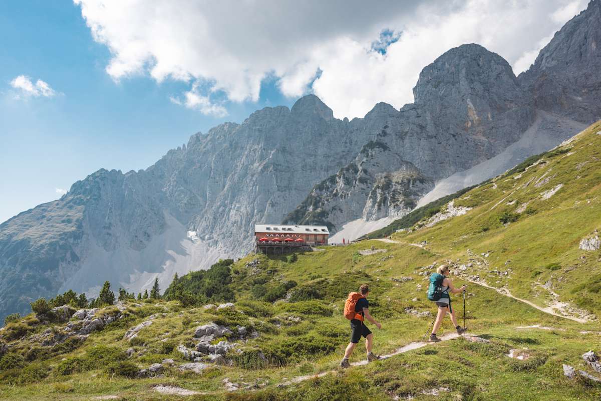 Zwei Wanderer nähern sich einer Alm, im Hintergrund steht eine Bergkette.