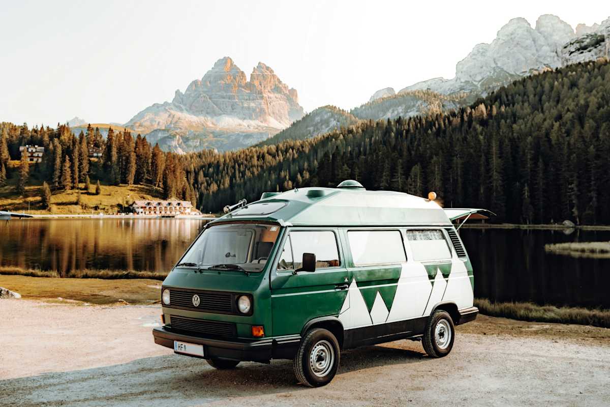Ein weiß-grüner Campervan steht vor einem See, im Hintergrund eine angeleuchtete Bergkulisse.