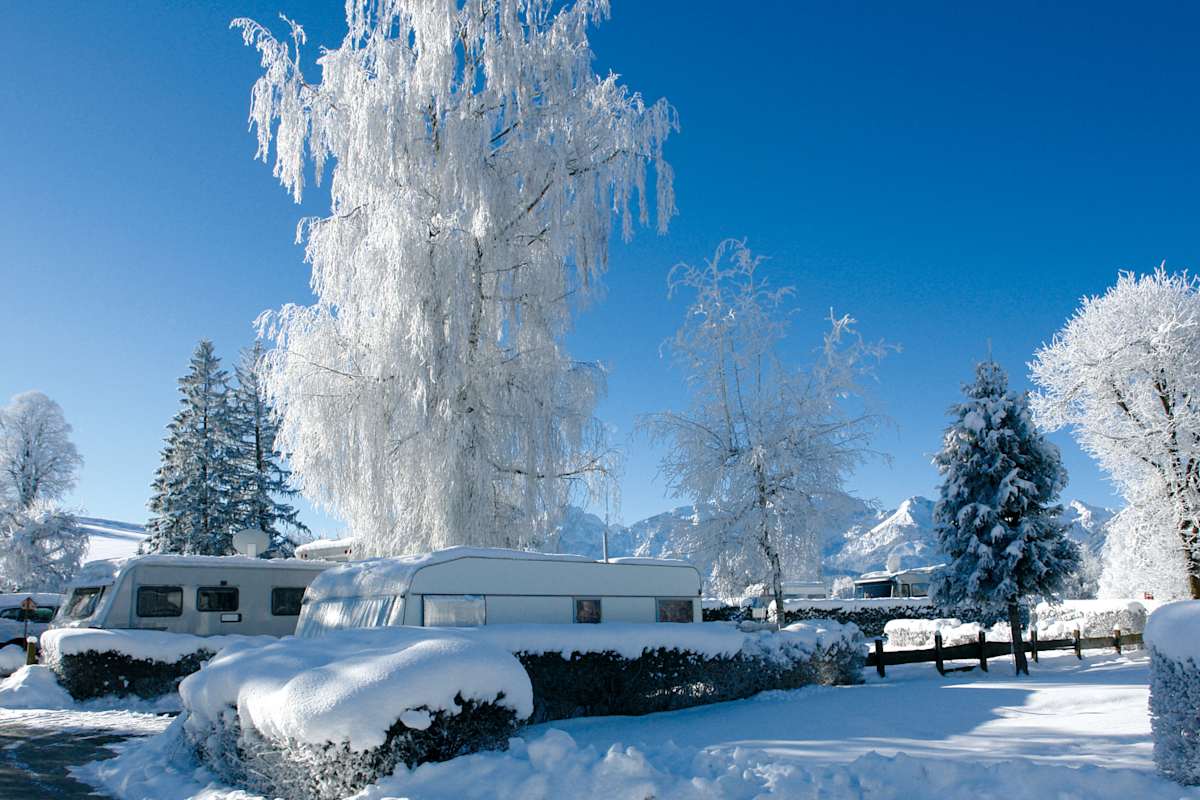 Der Campingplatz Hopfensee bei Füssen im Allgäu im Winter: Im Vordergrund eingeschneite Wohnwägen und Bäume, im Hintergrund eine Bergkulisse.