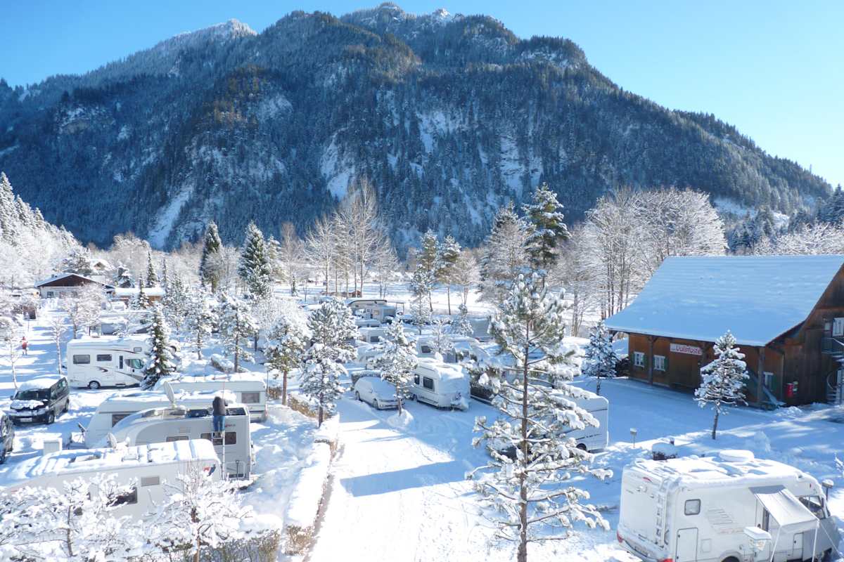 Blick auf den tief verschneiten Campingpark Oberammergau im Allgäu: Im Vordergrund verschneite Wohnwägen und Bäume, im Hintergrund eine bewaldete Bergkulisse.