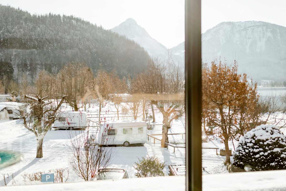 Blick von einem Haus auf den Camping Berau am Wolfgangsee. Im Vordergrund drei Campingwagen zwischen Bäumen, im Hintergrund eine Bergkulisse.