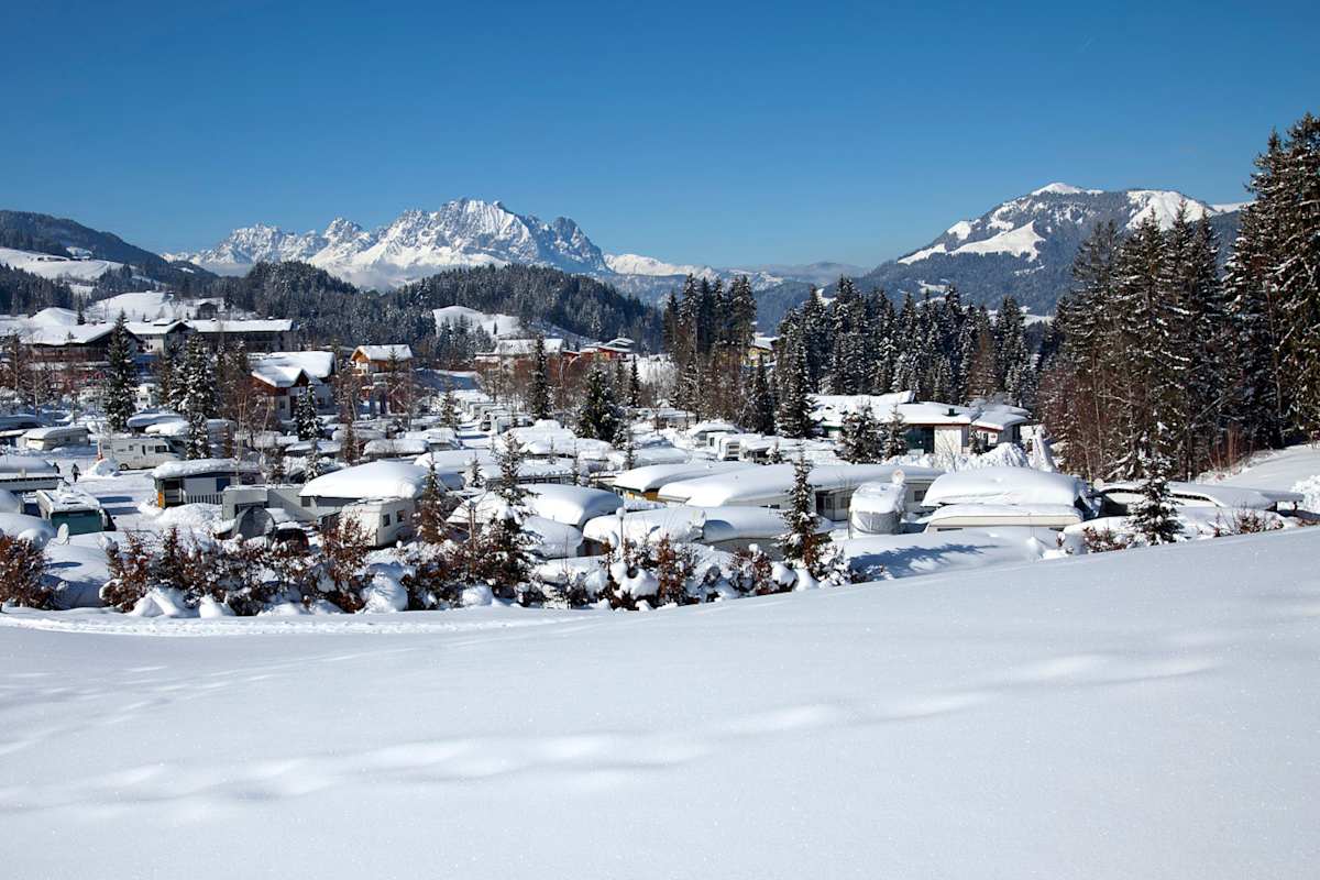 Blick in die tief verschneite Landschaft rund um das Tirol Camp in Fieberbrunn. Im Vordergrund die Wohnwägen, im Hintergrund eine verschneite Bergkulisse.