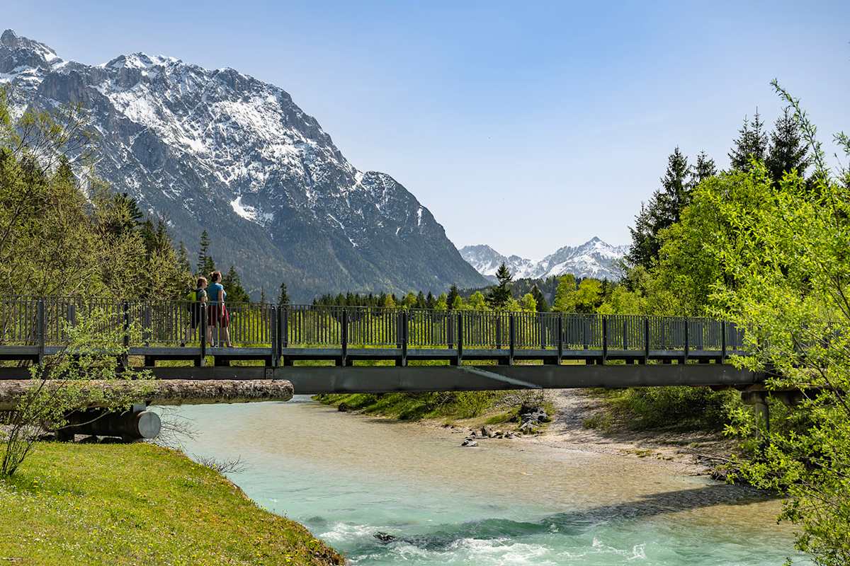 Zwei Frauen spazieren auf einer Brücke über die türkise Isar in einer sonnigen, bayerischen Landschaft.