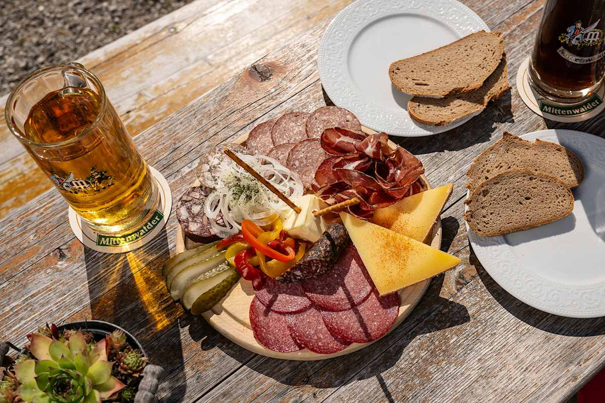 Zu sehen ist eine typisch bayerische Brettljause mit verschiedenen Speck- und Käsevariationen, Schwarzbrot und Mittenwalder Bier.