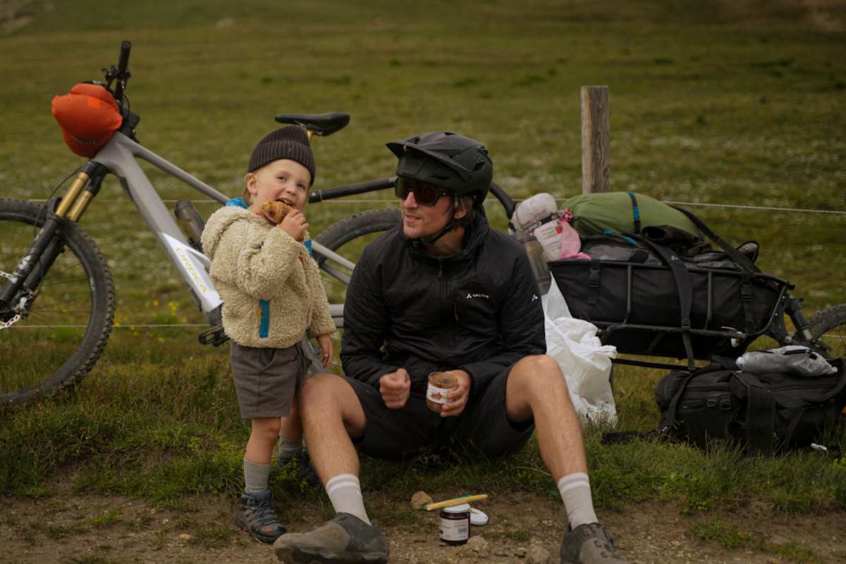 Ein Mann und ein Kind sitzen vor einem Fahrrad und essen zusammen eine Jause.