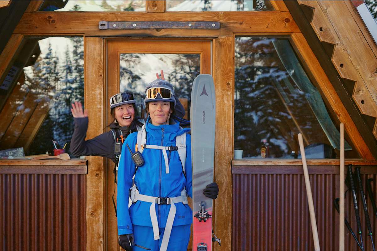 Zwei Frauen stehen vor einer Holzhütte mit ihrer Skiausrüstung und freuen sich.