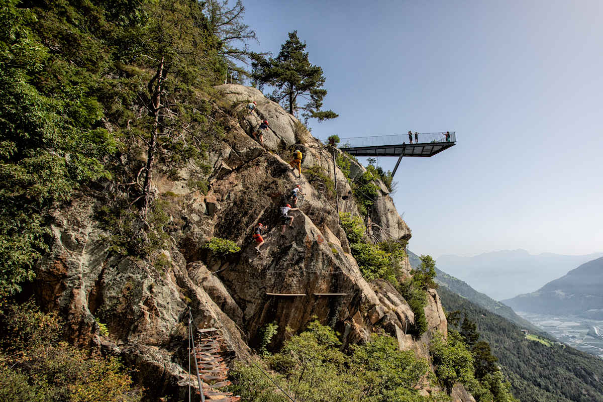 Kletterer klettern eine Bergwand in Naturns hoch, während ein paar andere Besucher auf einer Aussichtsplattform stehen.