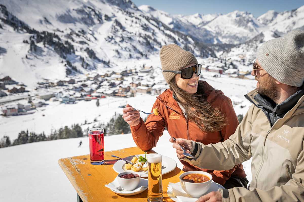 Eine Frau und ein Mann essen auf einer Hütte. Hinter ihnen ein ist ein Bergpanorama.