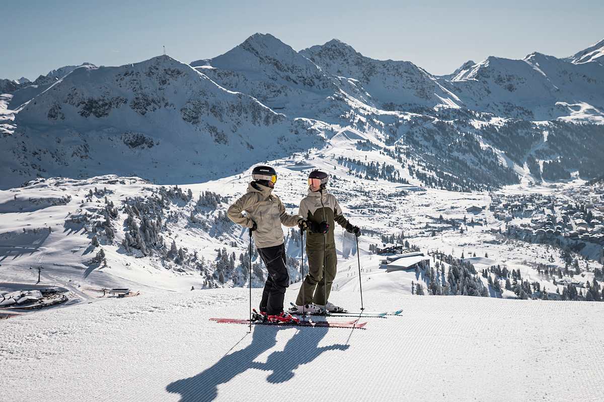 Eine Frau und ein Mann stehen auf einer perfekt präparierten Piste in Obertauern. Hinter ihnen ist ein schneereiches Bergpanorama zu sehen.