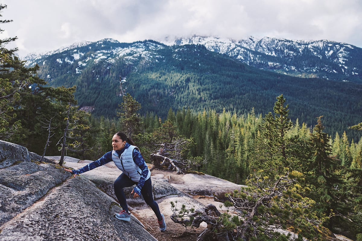 Eine Frau klettert in der Wildnis mit Outdoor-Bekleidung einen Hügel rauf.