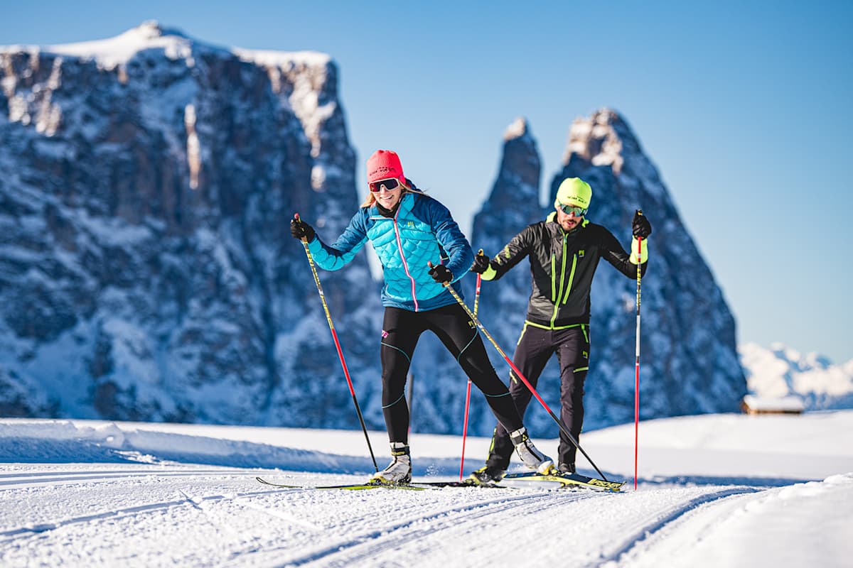 Ein eine Frau und ein Mann freuen sich über das Langlaufen auf einer der präparierten Loipen der Dolomitenregion Seiser Alm. Im Hintergrund sind markante Bergspitzen zu sehen.