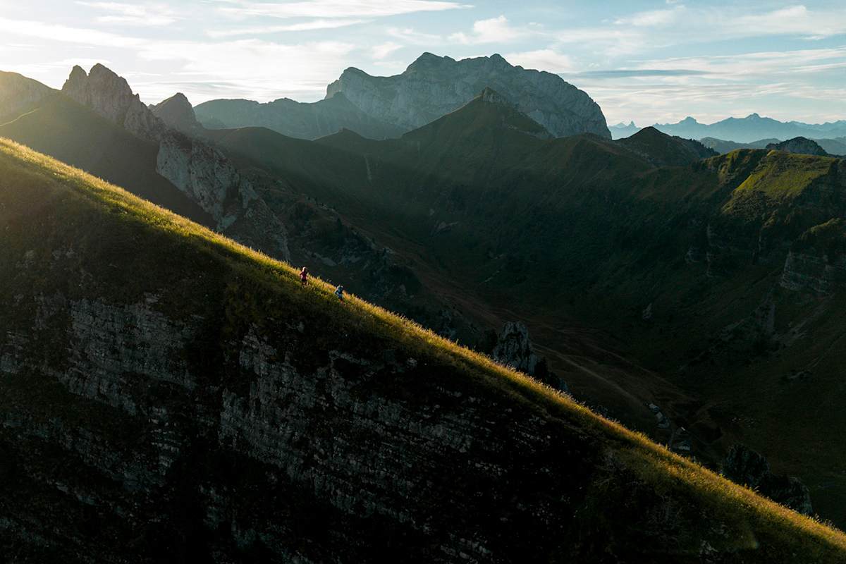 Man sieht eine beeindruckende, felsige Landschaft, in der zwei Personen einen Hang entlanglaufen. Ein Sonnenstrahl hebt die Laufstrecke besonders hervor.