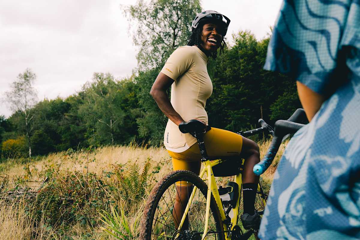 Eine Dame in gelben Radlershorts und Shirt von Endura sitzt auf einem Fahrrad und unterhält sich lachend mit einer anderen Person.