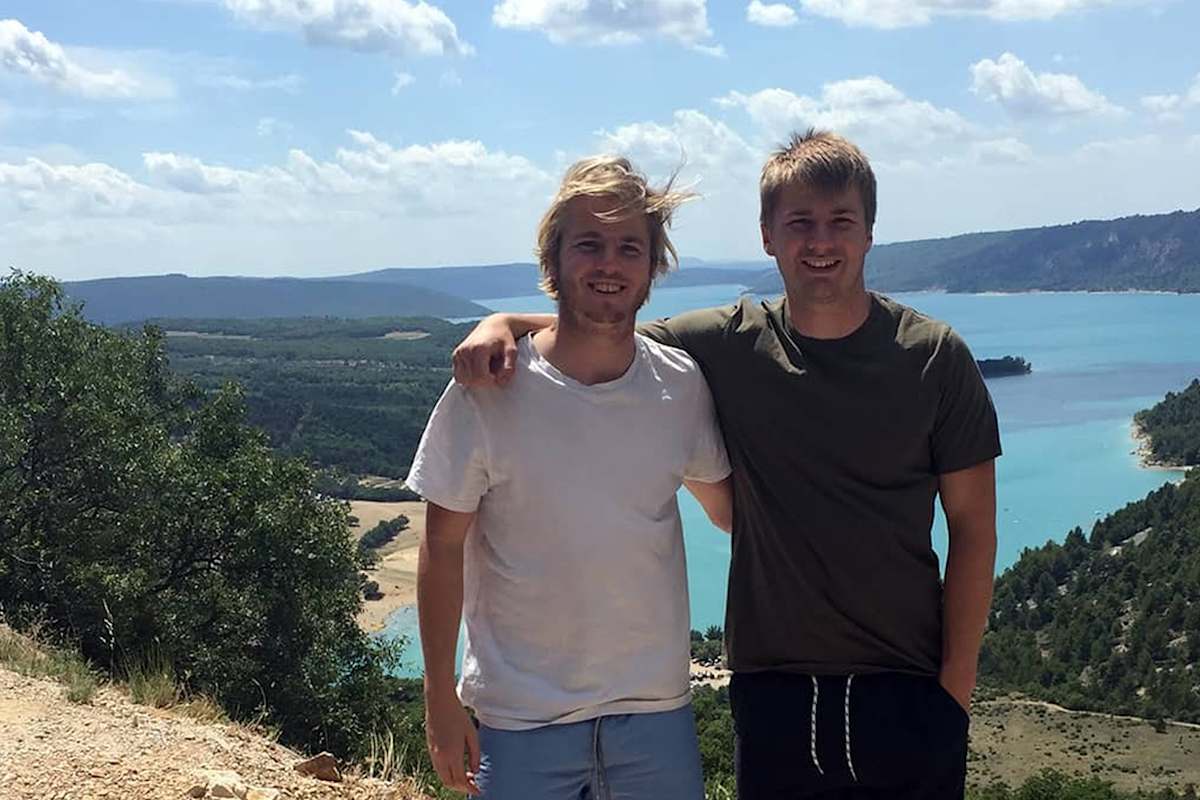 Zu sehen sind zwei Männer die sich lächelnd vor einer mediterranen Landschaft an einer Küste befinden.