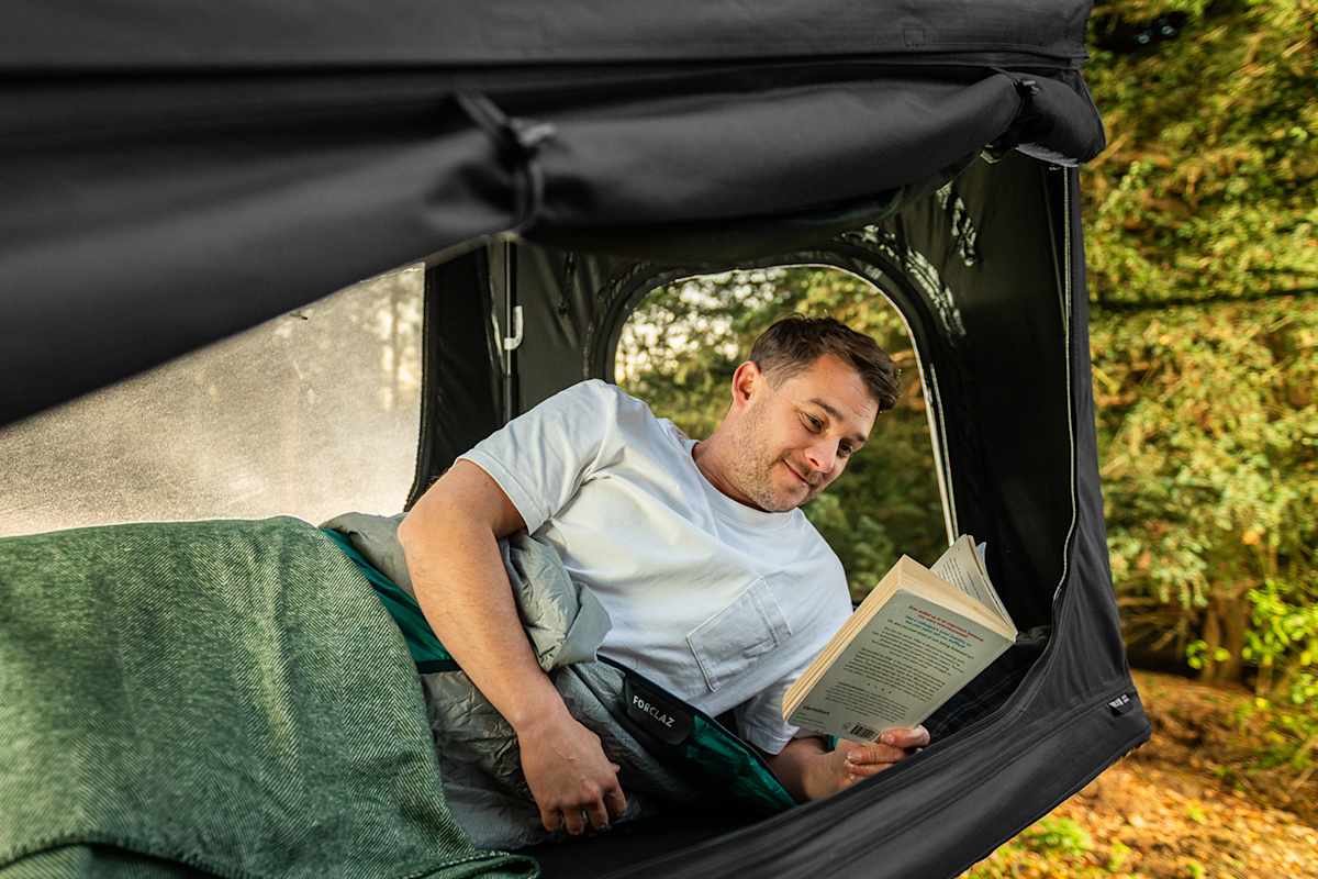 Ein Mann liegt im Inneren eines Dachzeltes und liest ein Buch bei offenem Fenster.