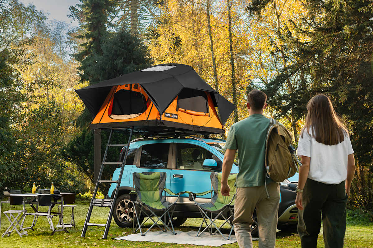 Ein Mann und eine Frau gehen auf ein Auto mit einem Dachzelt zu. Davor ist ein Campingbereich mit Tisch und Klappstühlen aufbereitet..