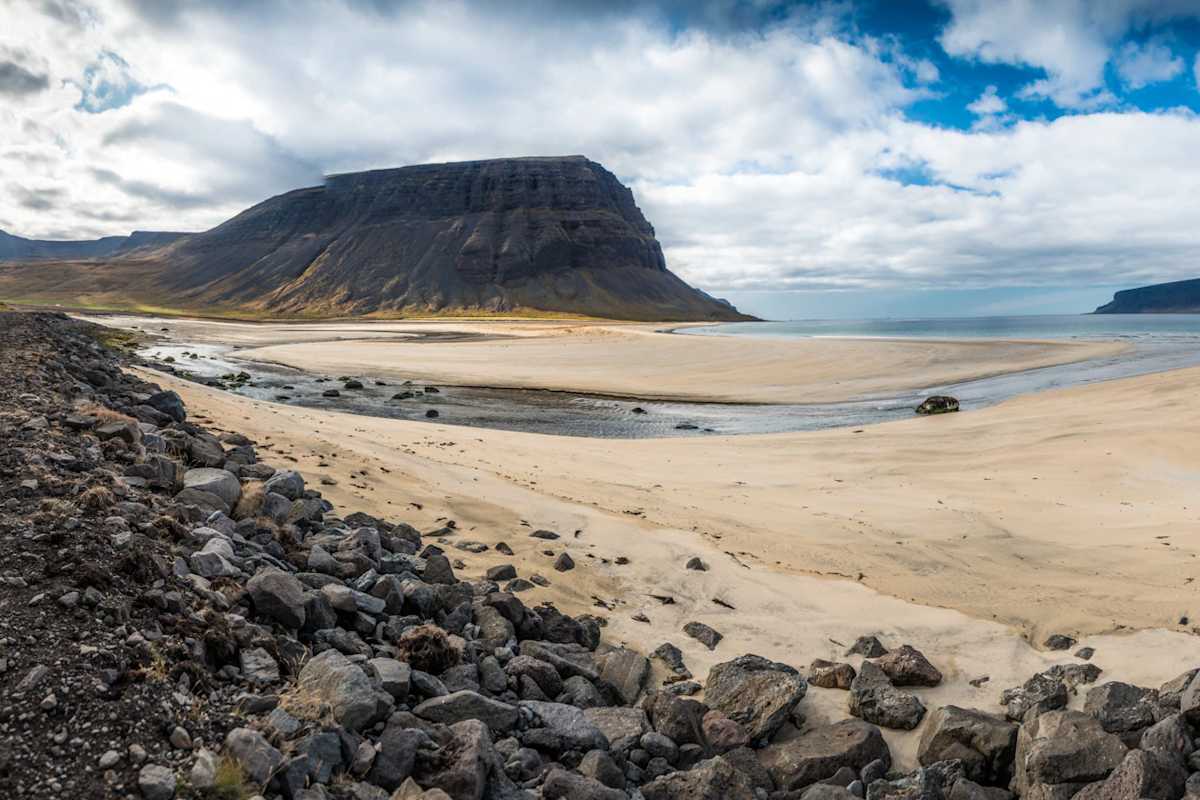 Heller Sandstrand mit dunklen Bergen und Meer im Hintergrund, im Vordergrund dunkles Gestein.