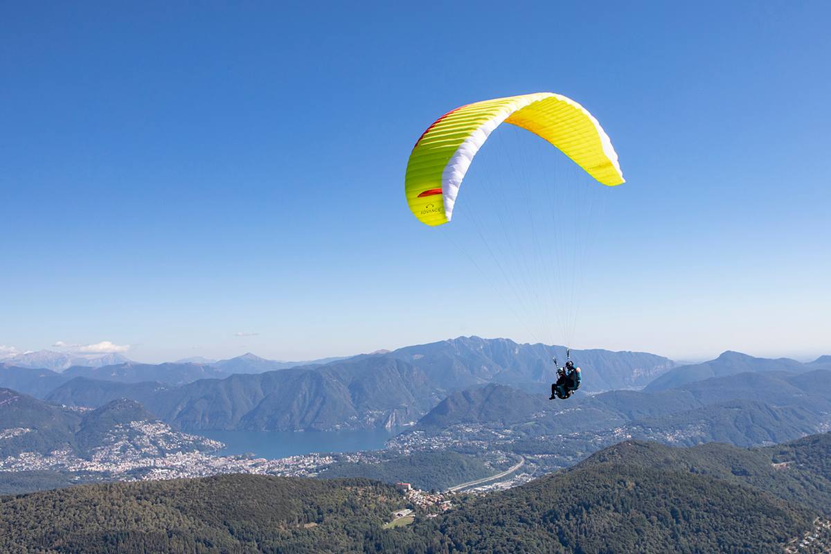 Man sieht zwei Personen die mit einem gelben Gleitschirm durch die Lüfte fliegen. Im Hintergrund sieht man eine wunderschöne, sonnige Landschaft mit grünen Hügeln und blauen Seen.