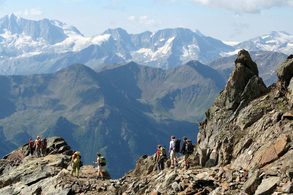 Wanderer überqueren einen steinigen Grat. Im Hintergrund schneebedeckte Berge.
