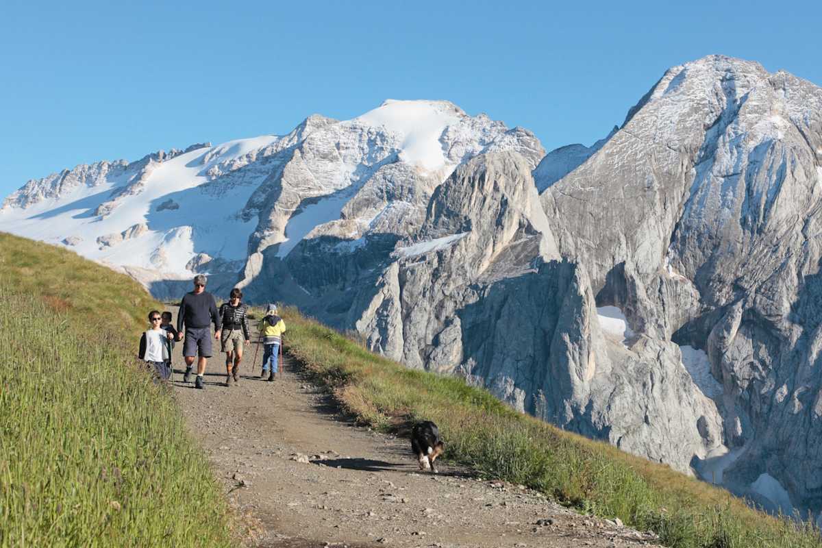 Eine Familie mit Hund wandert auf einem aussichtsreichen Höhenweg mit Blick auf die eisbedeckte Nordwand der Marmolata.