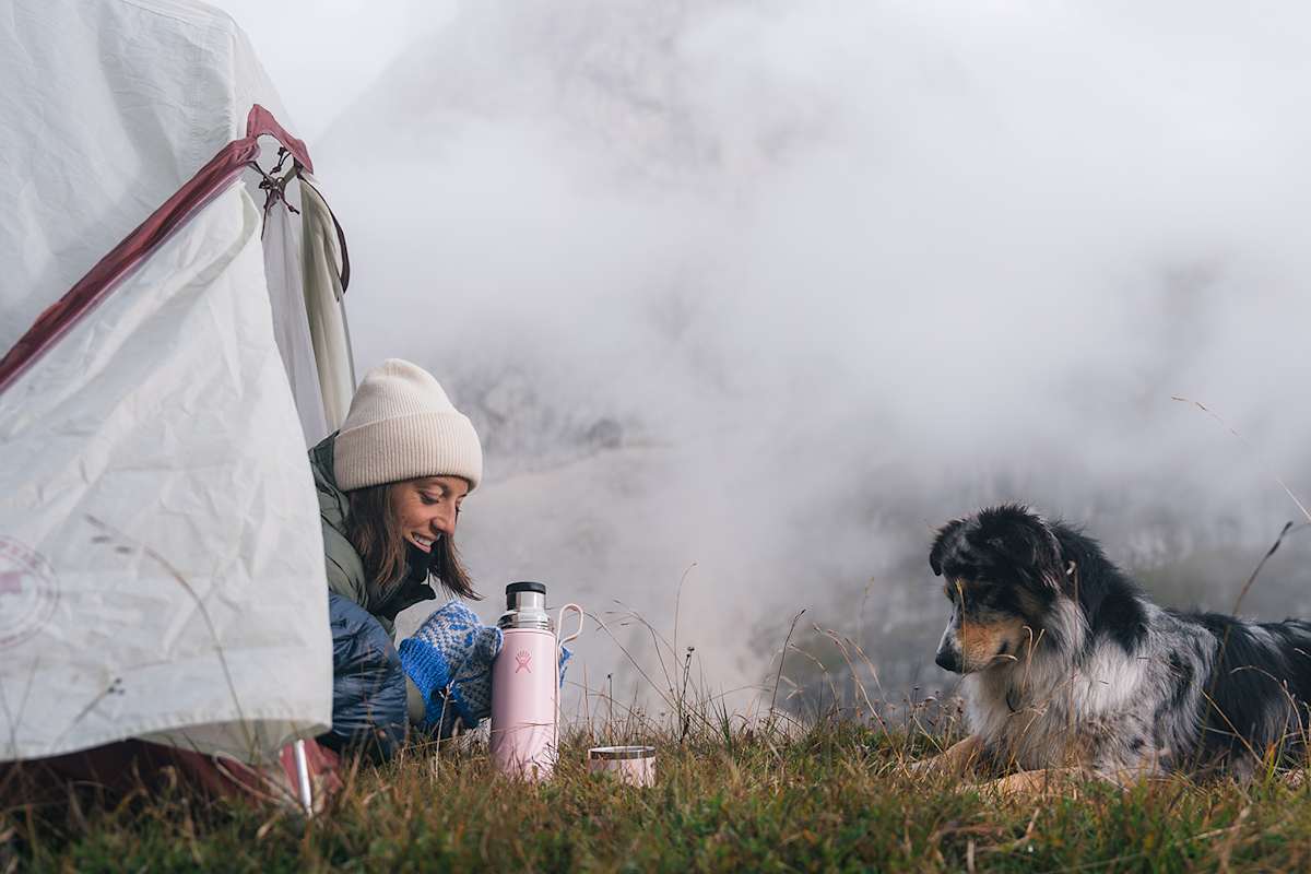 Frau im Zelt bei einer Bergwanderung mit Hund, umgeben von Nebel und Bergen, neben einer rosa Hydro Flask Hot Flask & Cup für heiße Getränke.