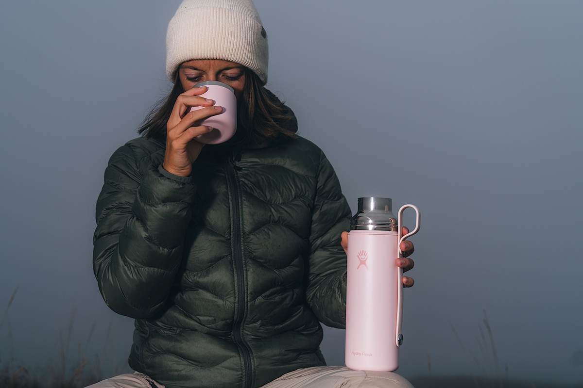 Frau in Winterkleidung genießt ein heißes Getränk aus der rosa Hydro Flask Hot Flask & Cup während einer Pause in nebliger Natur.
