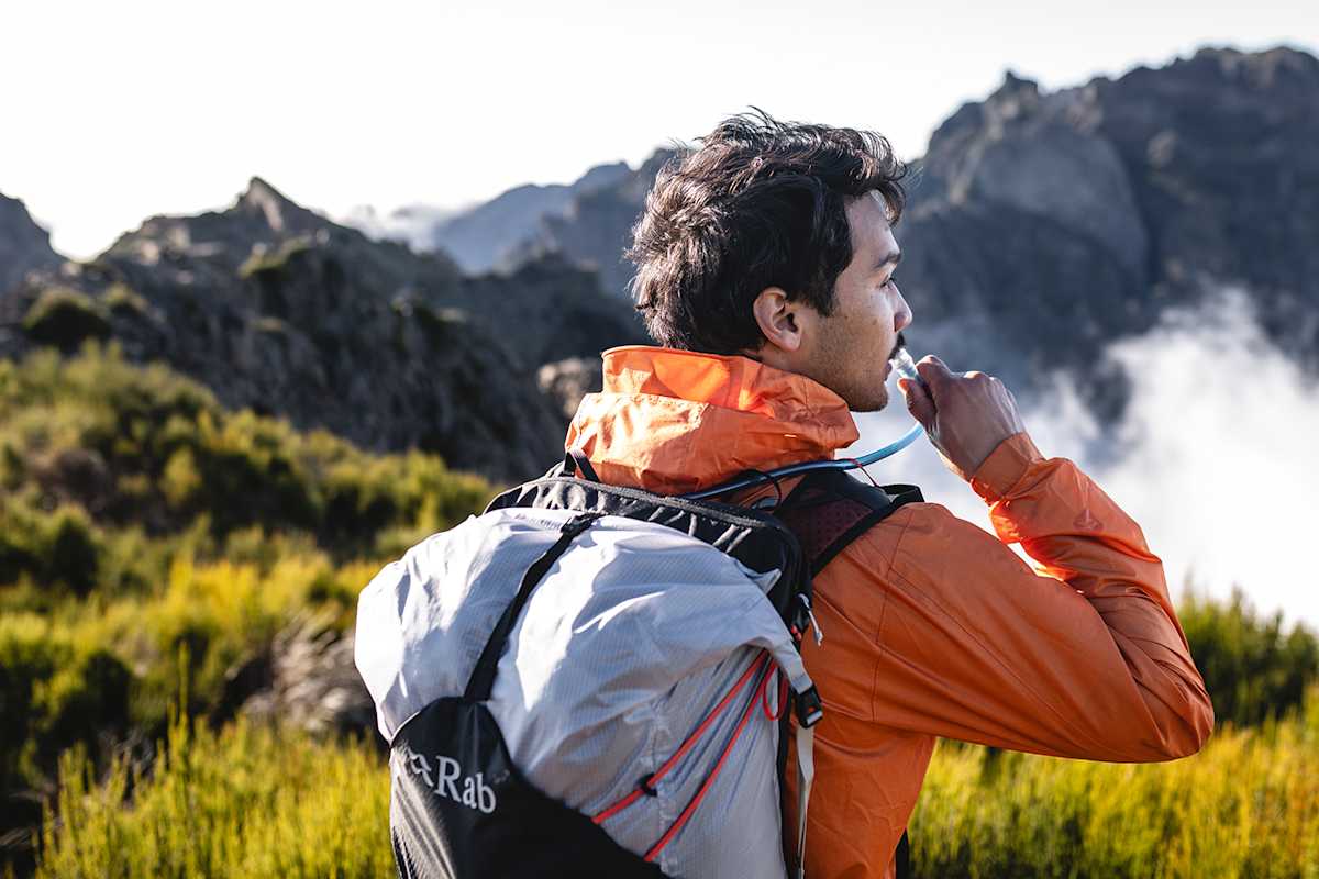 Ein Man trägt einen grauen Rab Rucksack und trinkt von einem Schlauch der aus dem Rucksack führt während er die Berglandschaft vor ihm genießt.