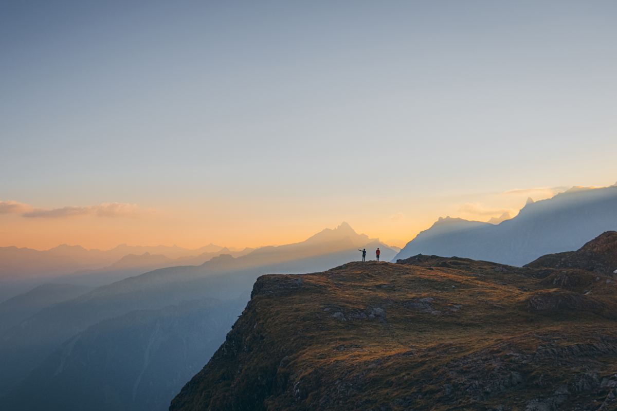 Kontrastreich: Layer für Layer präsentieren sich die Gipfel der Glarner Alpen im goldenen Licht der aufgehenden Sonne. Vor dem Horizont heben sich die zwei Personen am Berggipfel kontrastreich ab.