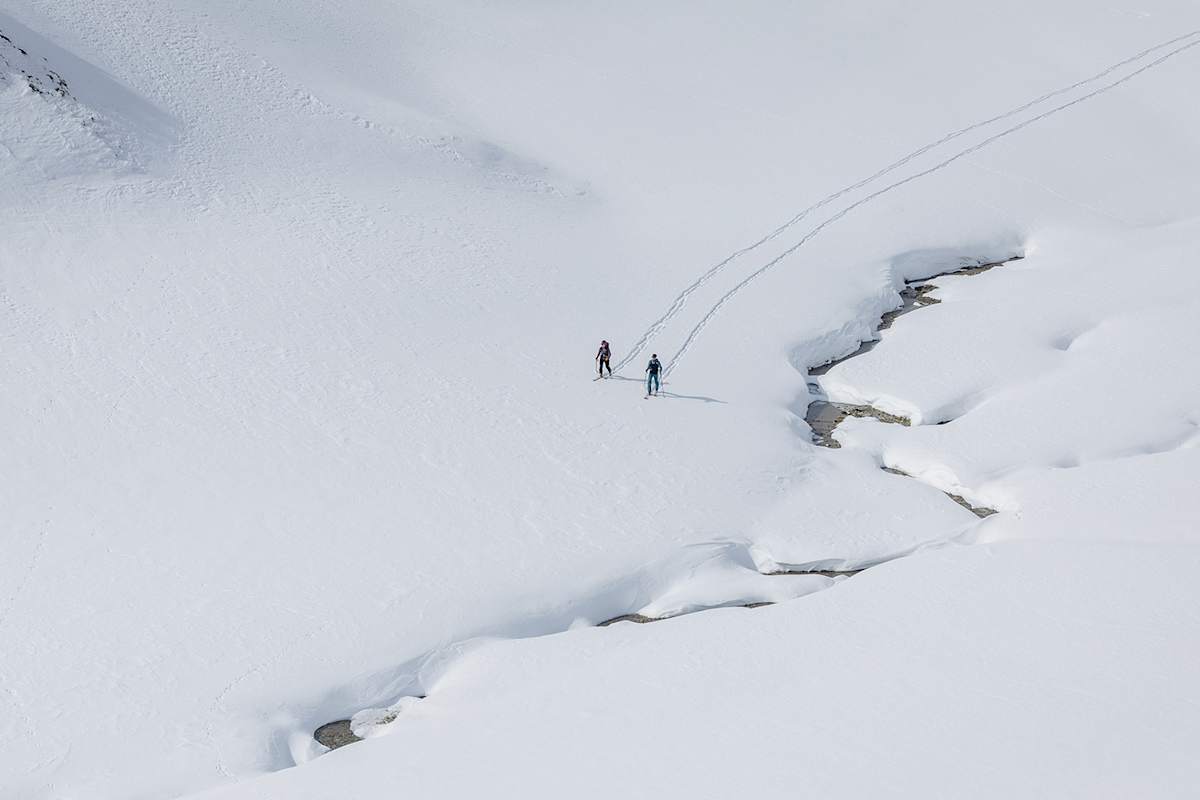 Zwei Skitourengeher durchqueren eine unberührte Schneelandschaft entlang eines Wasserlaufs.
