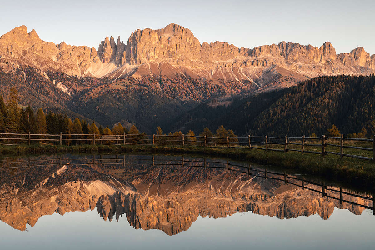 Zu sehen ist ein See inmitten einer alpinen Landschaft während des Sonnenuntergangs. Die Landschaft spiegelt sich im glasklaren Wasser sodass eine perfekte Reflektion entsteht.