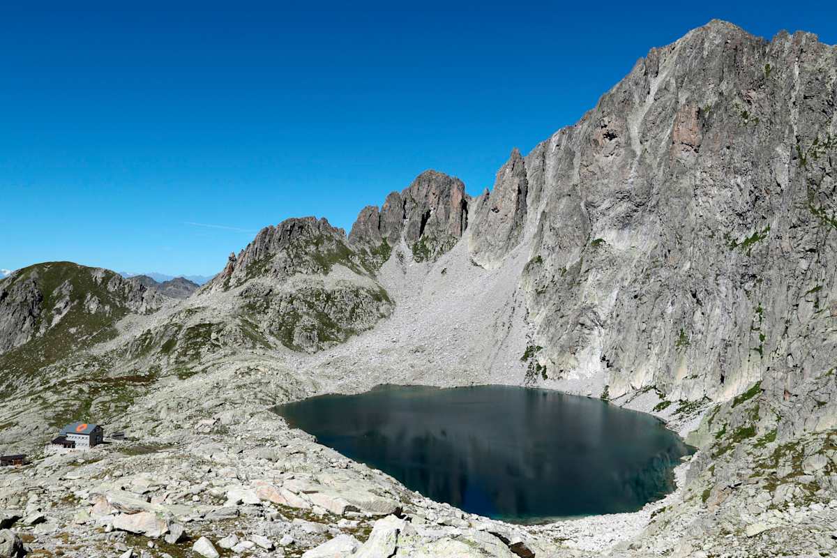 Ein grüner Bergsee vor einem rot-grauen Berggipfel. Links davon steht eine winzige Schutzhütte.