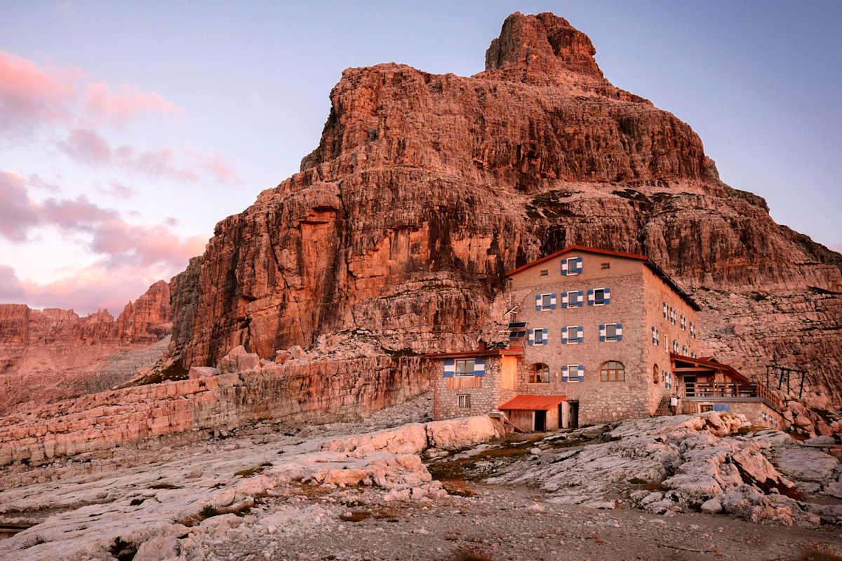 Eine Steinhütte mit blau-weißen Fensterläden am Fuße eines schroffen Dolomitengipfels im warmen Licht der aufgehenden Sonne.
