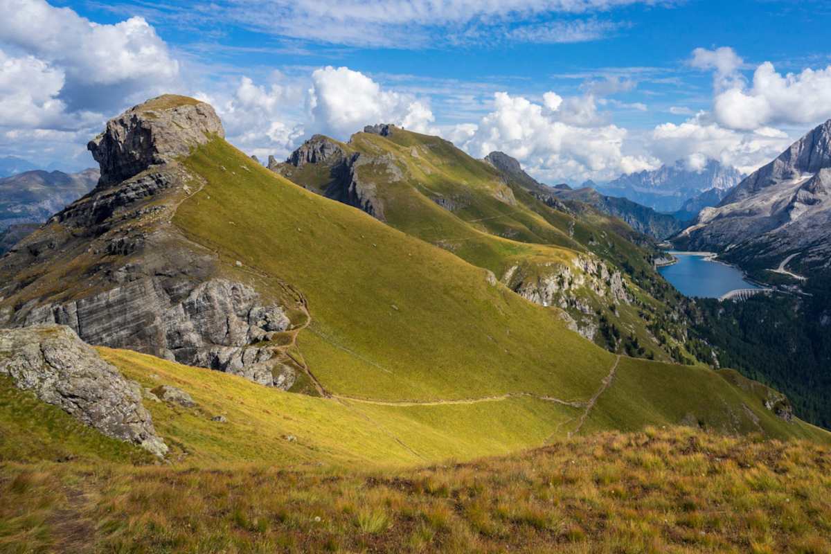 Im Vordergrund drei grasbewachsene Gipfel, im Hintergrund ein Stausee und die schroffen Felswände der Marmolada.