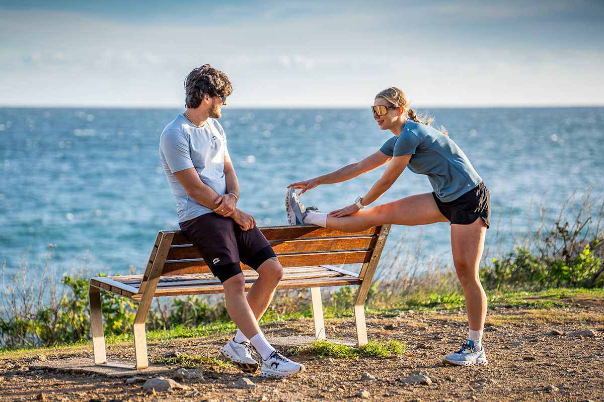 Eine Frau und ein Mann dehnen sich vor einer Bank. Sie tragen dabei Trailrunning-Outfits von skinfit. Hinter ihnen ist das Meer.