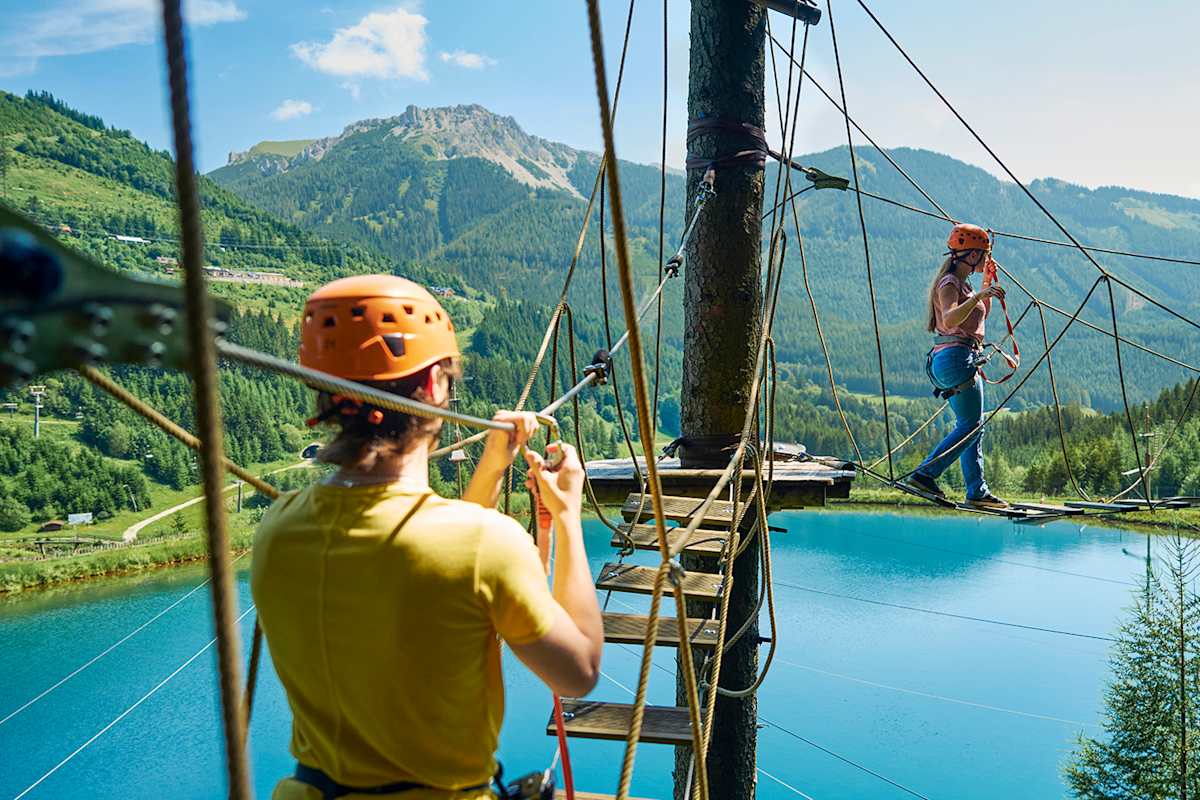 Zwei Personen im Hochseilgarten Alpfox überqueren eine Seilbrücke mit Blick auf den Grüblsee und die umliegenden Berge.