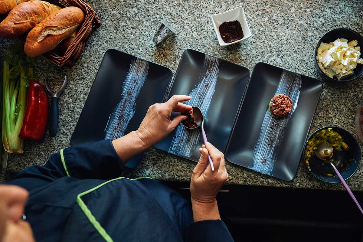 Eine Köchin richtet in der Seehütte am Grüblsee frisches Beef-Tatar auf schwarzen Tellern an, umgeben von Zutaten.