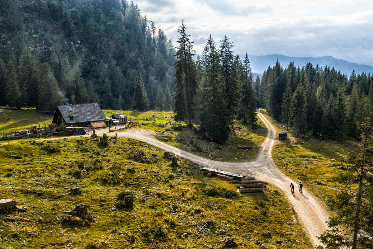 Zwei Gravelbiker fahren über eine Schotterstraße, im Hintergrund eine Almhütte.