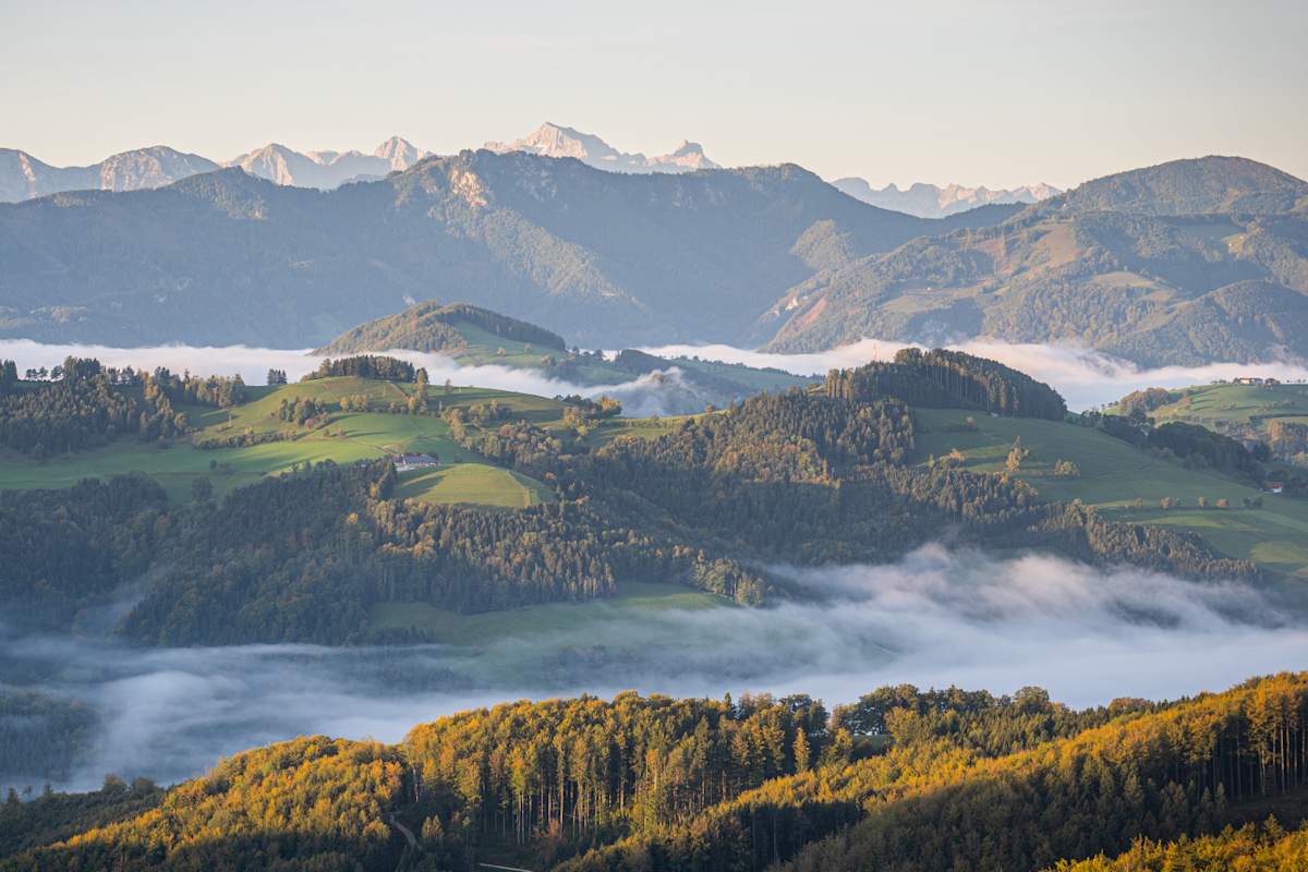 Eine hügelige Landschaft vom Sonnenlicht erleuchtet. In den Tälern wabern Nebelschwaden, im Hintergrund eine graue Bergkulisse.