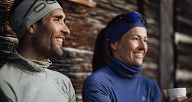 Zwei Personen in funktionaler Winterbekleidung von Odlo, genießen eine Pause im Freien vor einer rustikalen Holzhütte.