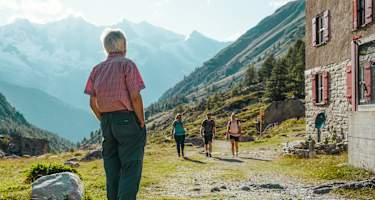 Ein Mann steht vor einer Hütte und wartet auf zwei Wanderinnen und einen Wanderer.
