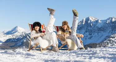 Zwei Frauen sitzen auf Liegestühlen auf einem Berg oben und heben jeweils einen Fuß in die Höhe.