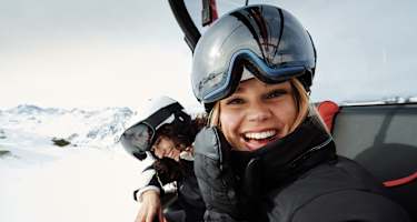 Eine Frau mit Skibrille und Helm auf dem Kopf sitz in einem Sessellift und lacht uns entgegen. Neben ihr sitzt eine weitere lächelnde Frau.