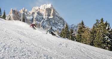 Zwei Skifahren carven in hohem Tempo über eine Skipiste. Im Hintergrund ein schroffer Dolomitengipfel