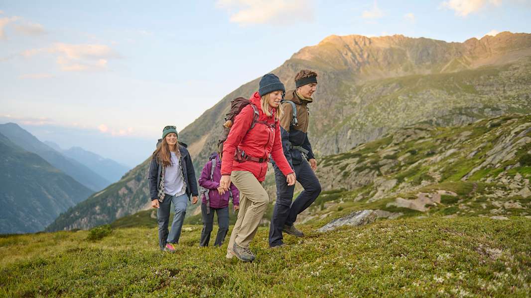Eine Gruppe von Wanderern in funktionaler Outdoor-Bekleidung bewegt sich durch eine malerische Berglandschaft. Grüne Wiesen und majestätische Gipfel bilden eine beeindruckende Kulisse.